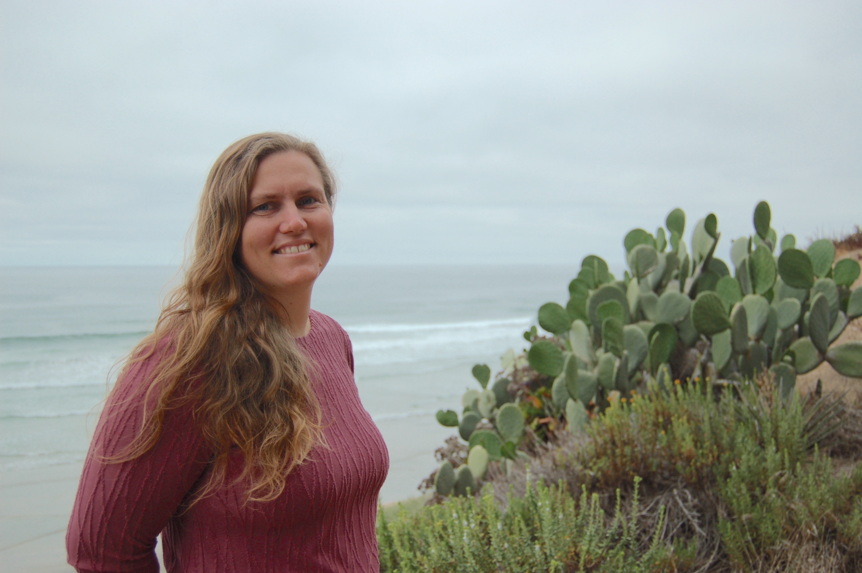 Jessica Goodheart standing on a beach by the ocean next to cactus plants.