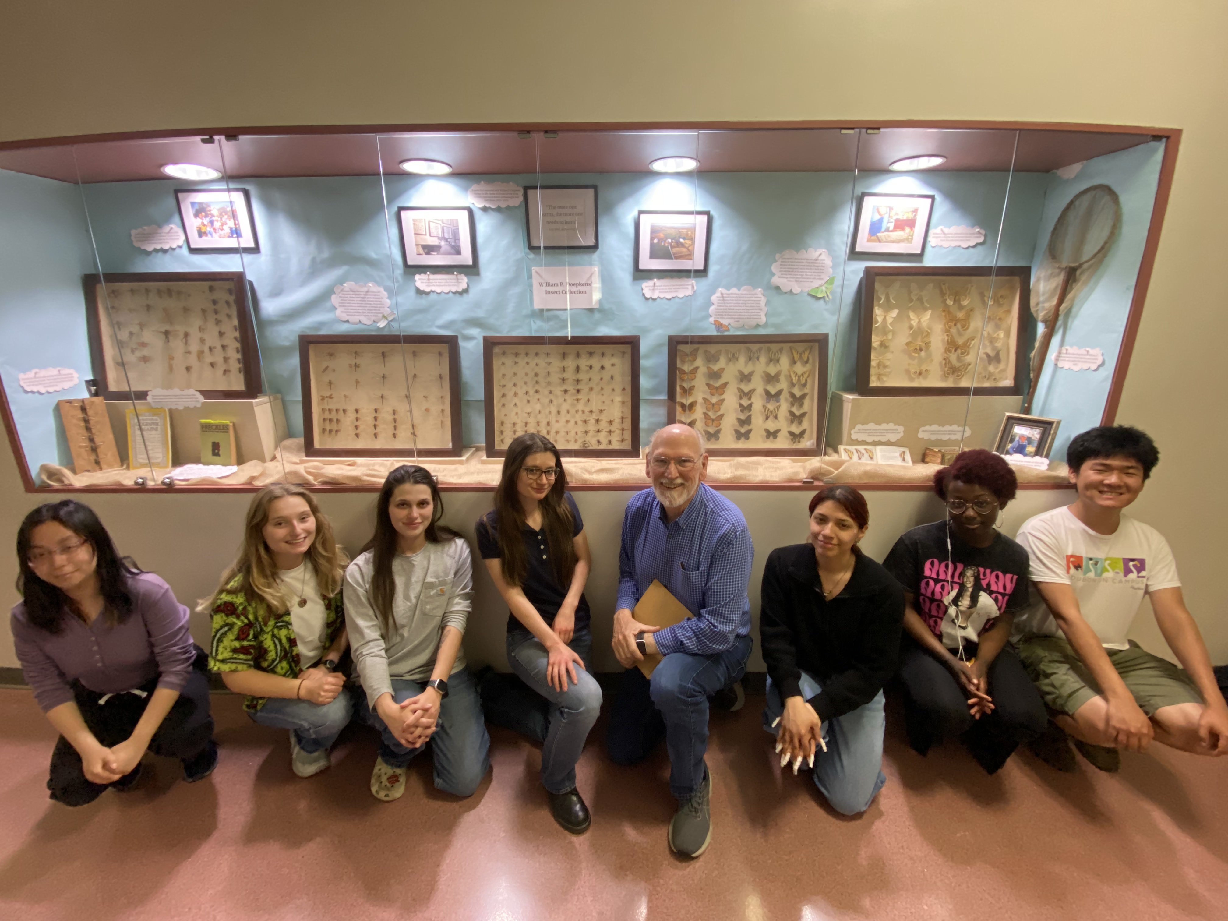Students and instructors smiling for photo in front of insect collection on  display in hallway cabinet