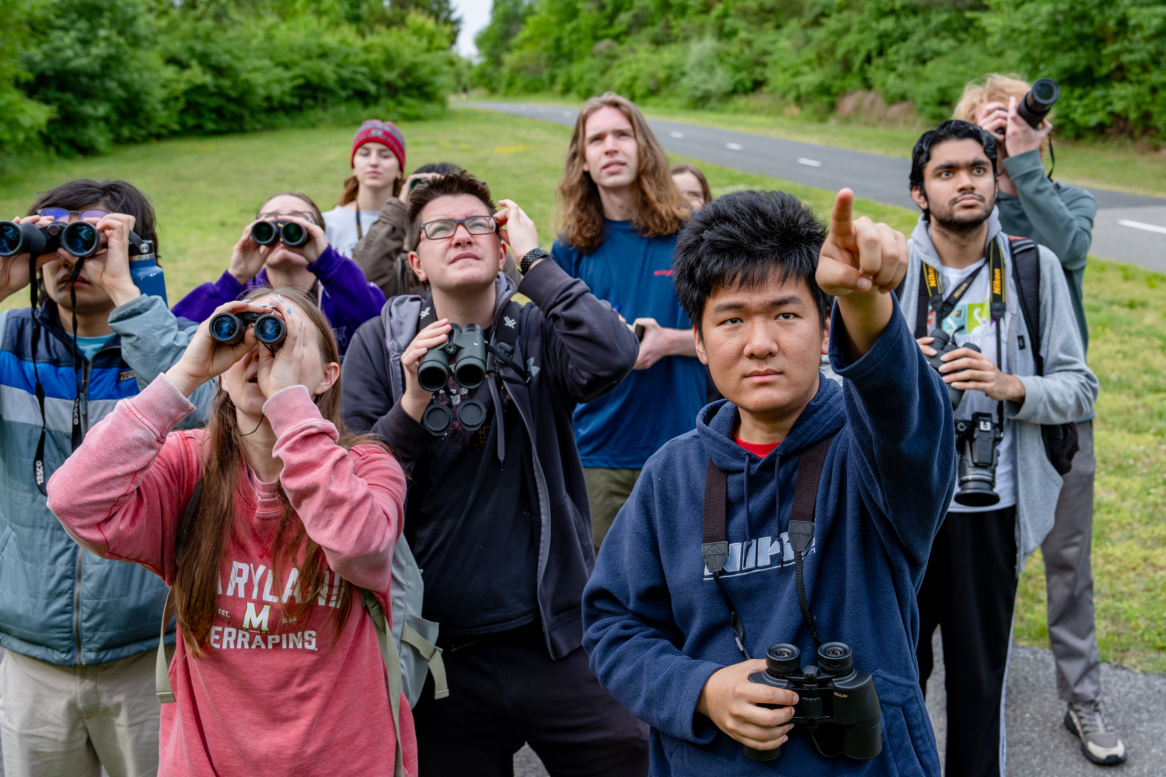 A man in a blue hoodie points as nine students behind him look through binoculars. 