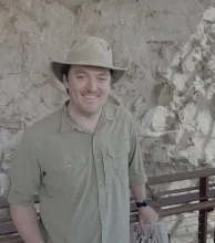 Nathan Jud in brown hat and shirt smiles while standing in front of a rock wall.