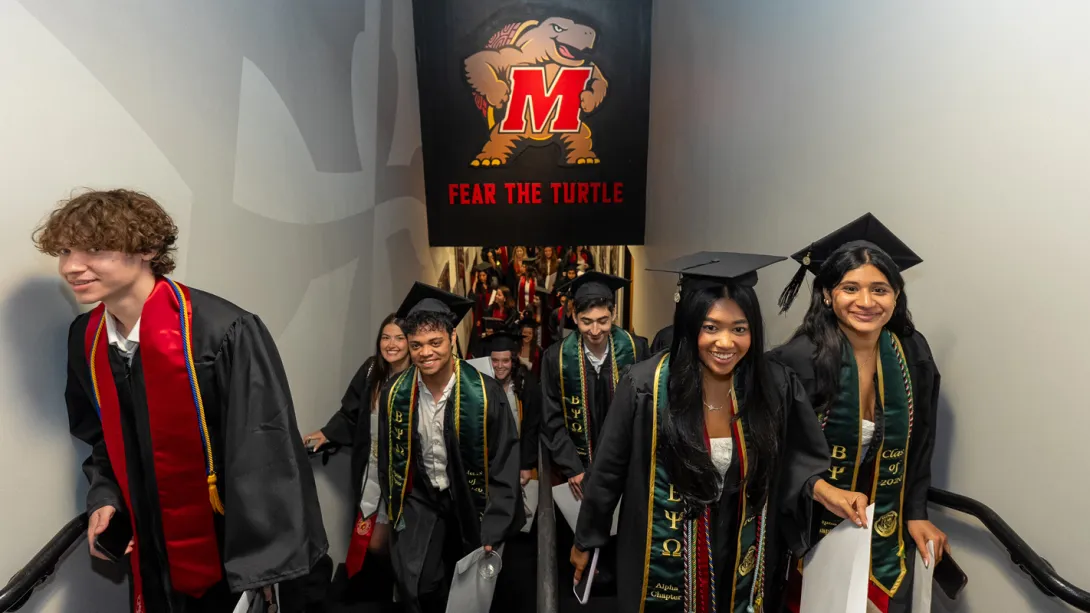 Graduates coming up stairs under a picture of Testudo, on their way to the graduation ceremony.